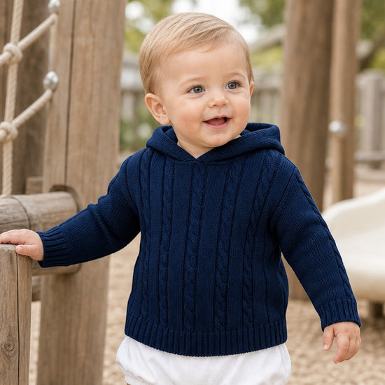 Carriage Boutique navy cable knit hooded baby sweater worn by a smiling toddler in a playground