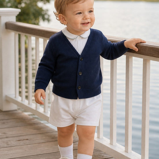 Carriage Boutique navy cotton V-neck cardigan worn by a toddler boy with white shorts and shoes on a wooden deck