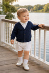 Carriage Boutique navy cotton V-neck cardigan worn by a toddler boy with white shorts and shoes on a wooden deck