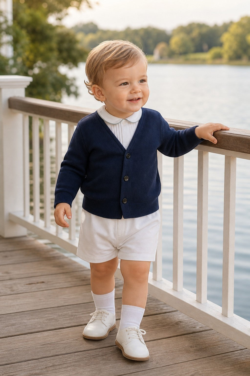 Carriage Boutique navy cotton V-neck cardigan worn by a toddler boy with white shorts and shoes on a wooden deck