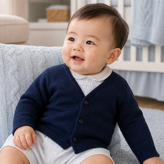 Baby boy wearing a Carriage Boutique navy cotton V-neck cardigan over a white collared shirt in a nursery setting.