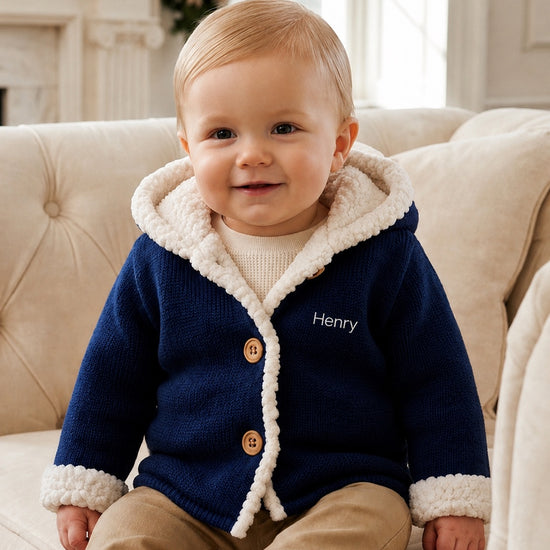 Carriage Boutique navy knit cotton baby jacket with white sherpa lining, hood, and front buttons, worn by a toddler.