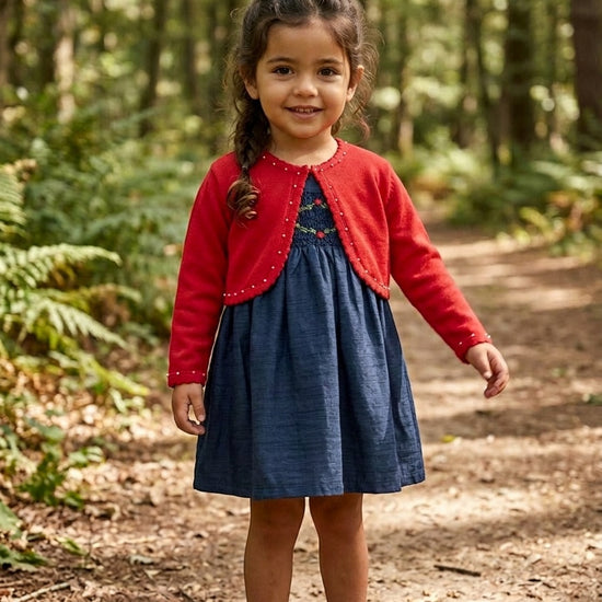 Carriage Boutique navy silk floral smocked dress with a red cardigan worn by a young girl on a forest path