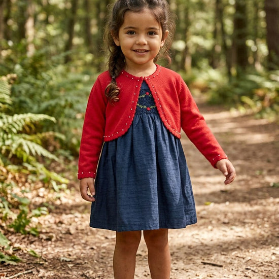 Carriage Boutique navy silk dress with smocked floral details and red cardigan worn by a girl in a forest setting