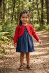 Carriage Boutique navy silk dress with smocked floral details and red cardigan worn by a girl in a forest setting