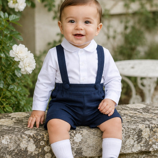 Carriage Boutique baby boy navy poly silk suspender shorts set with a white long sleeve shirt sitting on a stone wall