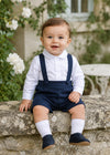 Carriage Boutique baby boy navy poly silk suspender shorts set with a white long sleeve shirt sitting on a stone wall