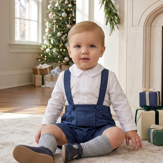 Carriage Boutique baby boy wearing a navy poly silk suspender shorts set with a white long-sleeve shirt by a tree.
