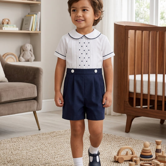 Carriage Boutique toddler boy wearing a white shirt with navy tucks and dots paired with navy blue pleated shorts.
