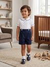 Carriage Boutique toddler boy wearing a white shirt with navy tucks and dots paired with navy blue pleated shorts.