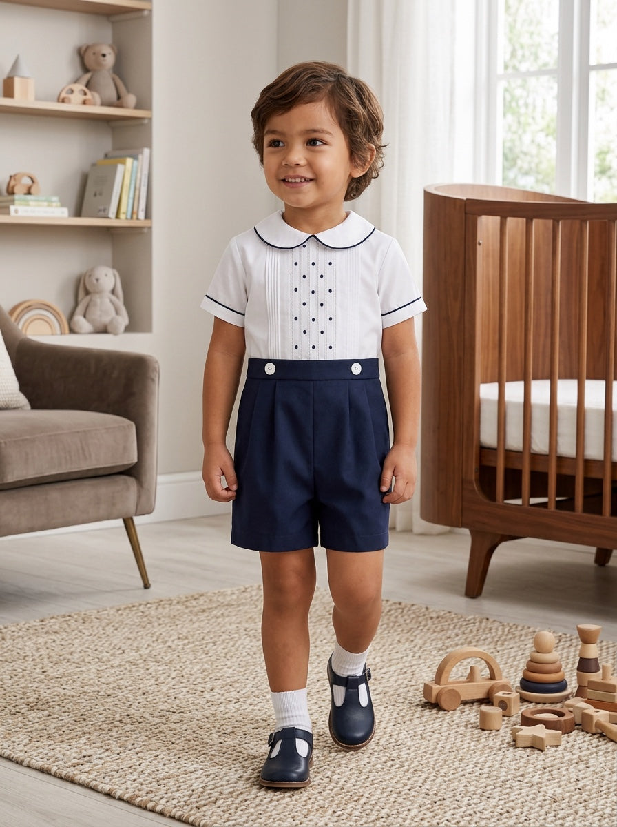 Carriage Boutique toddler boy wearing a white shirt with navy tucks and dots paired with navy blue pleated shorts.