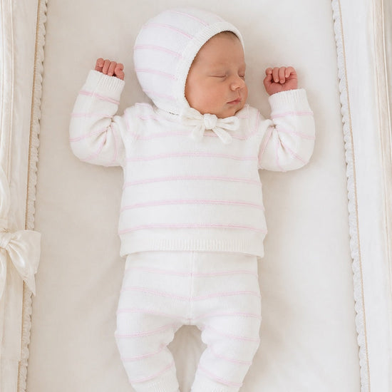 Newborn baby sleeping in a Carriage Boutique white knit set with blush pink stripes, including leggings and a bonnet.