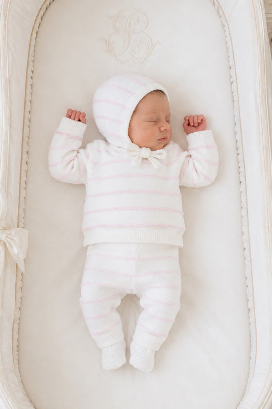 Newborn baby sleeping in a Carriage Boutique white knit set with blush pink stripes, including leggings and a bonnet.
