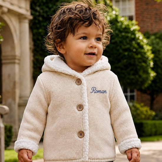Carriage Boutique off-white knit cotton hooded jacket with sherpa lining and blue monogram on a toddler outdoors