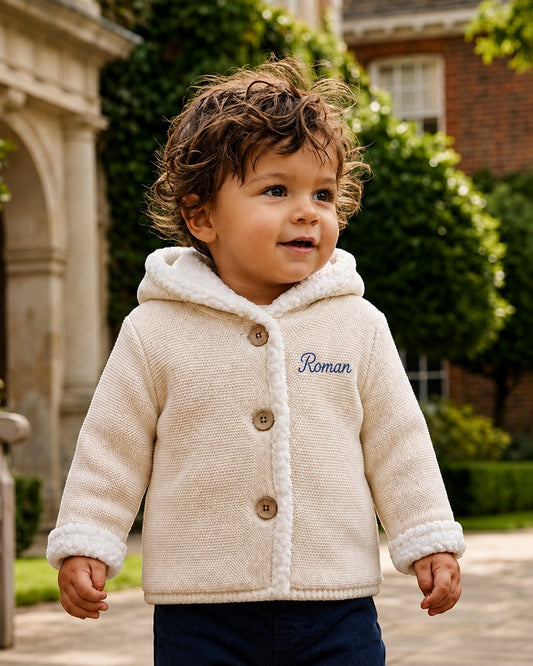 Carriage Boutique off-white knit cotton hooded jacket with sherpa lining and blue monogram on a toddler outdoors
