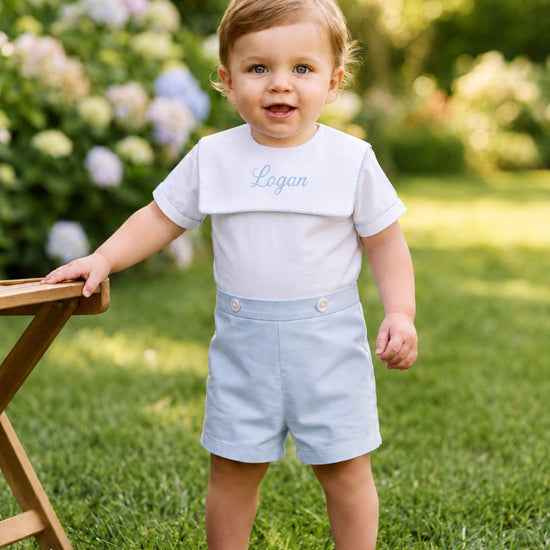 Carriage Boutique personalized white and light blue cotton bobby suit worn by a toddler in a garden setting.