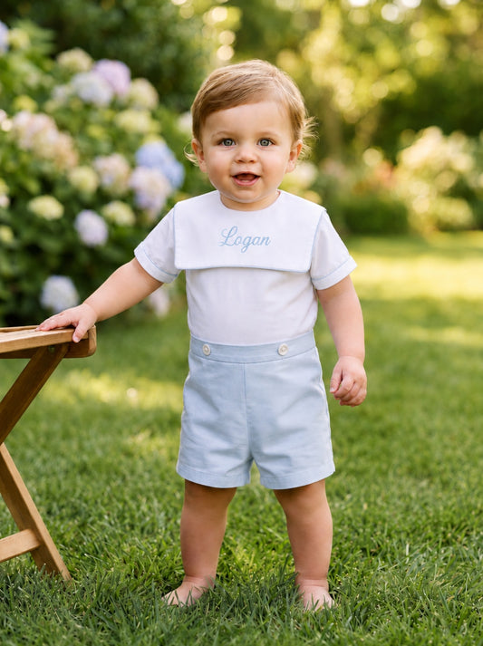Carriage Boutique personalized white and light blue cotton bobby suit worn by a toddler in a garden setting.