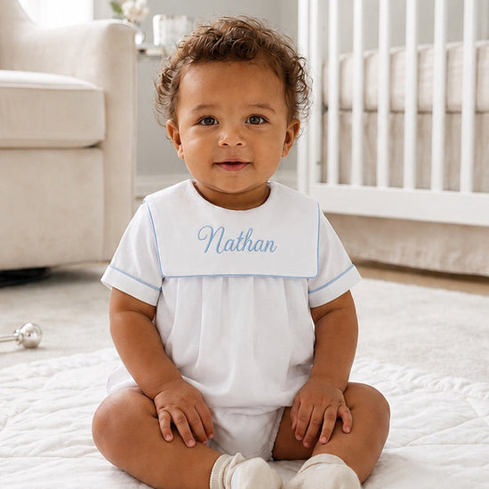 Baby boy wearing a Carriage Boutique white cotton romper with blue trim and personalized Nathan embroidery