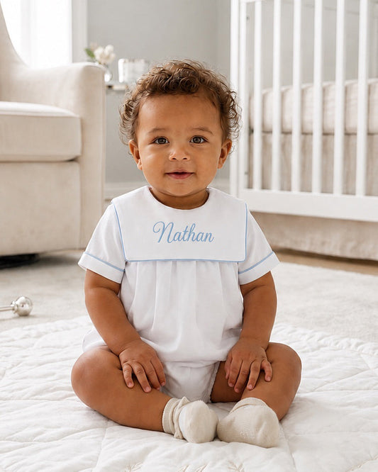 Baby boy wearing a Carriage Boutique white cotton romper with blue trim and personalized Nathan embroidery