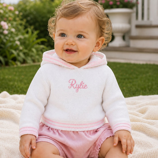 Carriage Boutique white hooded sweater with pink trim and personalized embroidery worn by a baby girl outdoors.