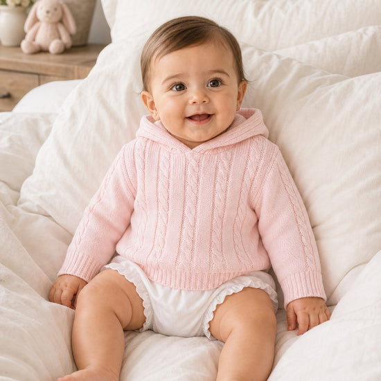 Carriage Boutique baby wearing a soft pink cotton cable knit hooded sweater while sitting on a white bed.