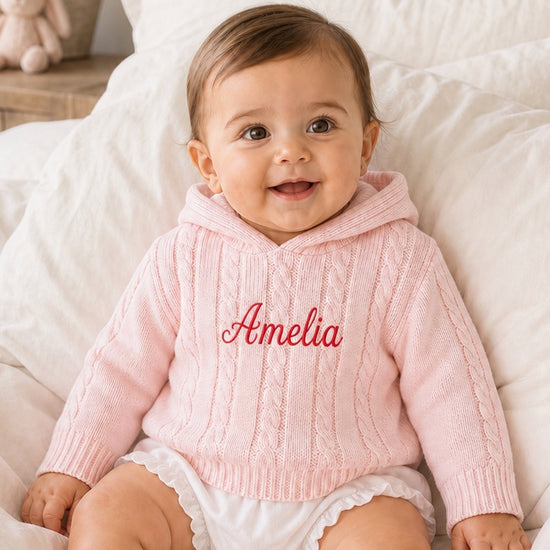 Carriage Boutique personalized pink cable knit baby sweater with hood and embroidered name on white bedding