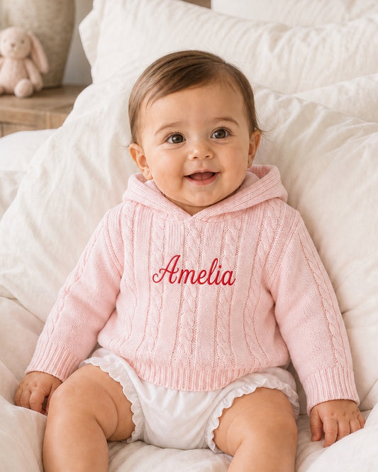 Carriage Boutique personalized pink cable knit baby sweater with hood and embroidered name on white bedding