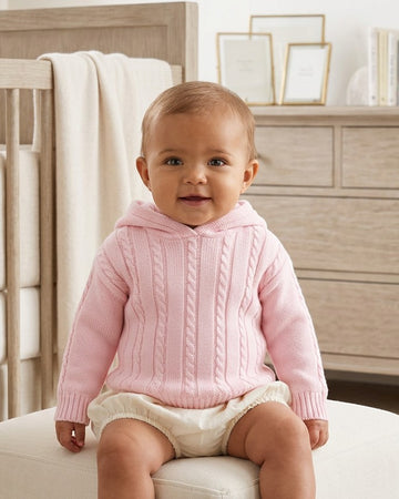 Carriage Boutique baby wearing a soft pink cable knit cotton hooded sweater with a zip-back design in a nursery.