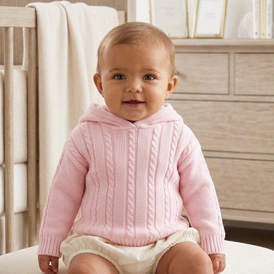 Baby wearing a Carriage Boutique pink cable knit hooded cotton sweater with a zip-back design in a nursery.