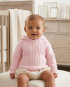 Baby wearing a Carriage Boutique pink cable knit hooded cotton sweater with a zip-back design in a nursery.