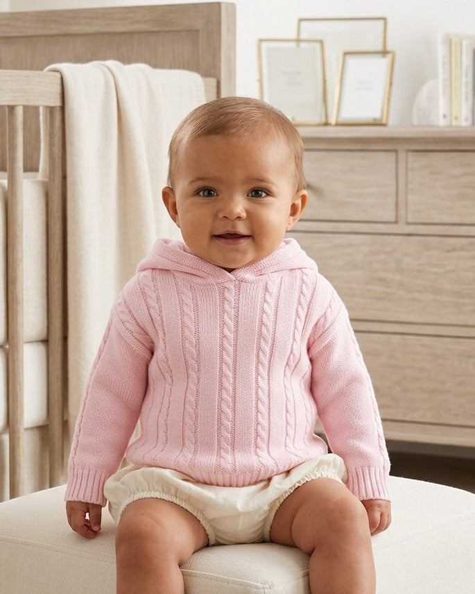 Baby wearing a Carriage Boutique pink cable knit hooded cotton sweater with a zip-back design in a nursery.
