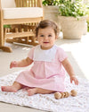Baby girl wearing a Carriage Boutique personalized pink and white cotton dress with a button bib, sitting on a blanket
