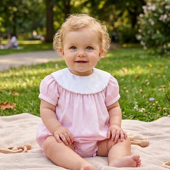Carriage Boutique baby girl wearing a light pink cotton bubble romper with a white Peter Pan collar in a park.