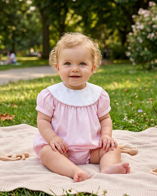 Carriage Boutique baby girl wearing a light pink cotton bubble romper with a white Peter Pan collar in a park.