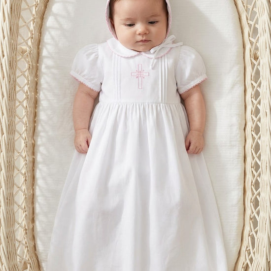 Carriage Boutique baby girl christening gown in white with pink embroidered cross and matching bonnet in a wicker bassinet