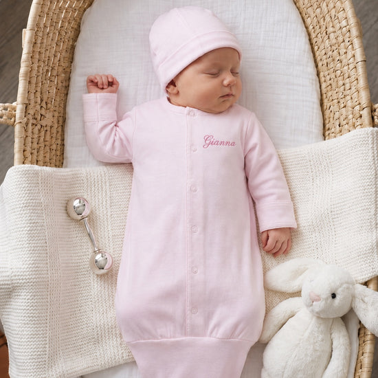 Carriage Boutique pink newborn sleep sack with matching hat and personalized name embroidery in a wicker bassinet.