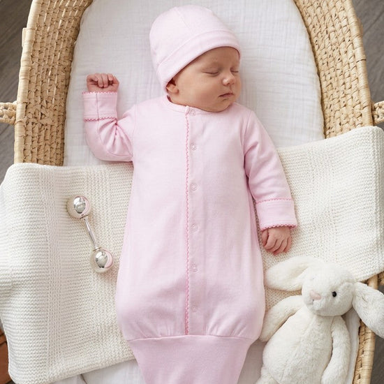 Carriage Boutique newborn pink sleep sack with matching hat, featuring scalloped trim and knotted bottom in a bassinet.