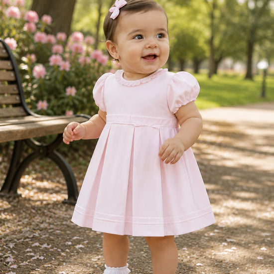 Carriage Boutique baby girl wearing a pink pique dress with puff sleeves and a matching bow headband in a park.