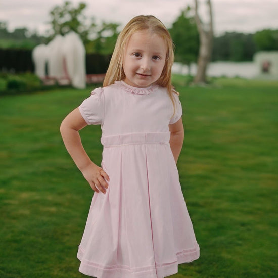 Carriage Boutique pink pique baby girl dress with ruffled collar, worn by a young girl in a grassy outdoor setting.
