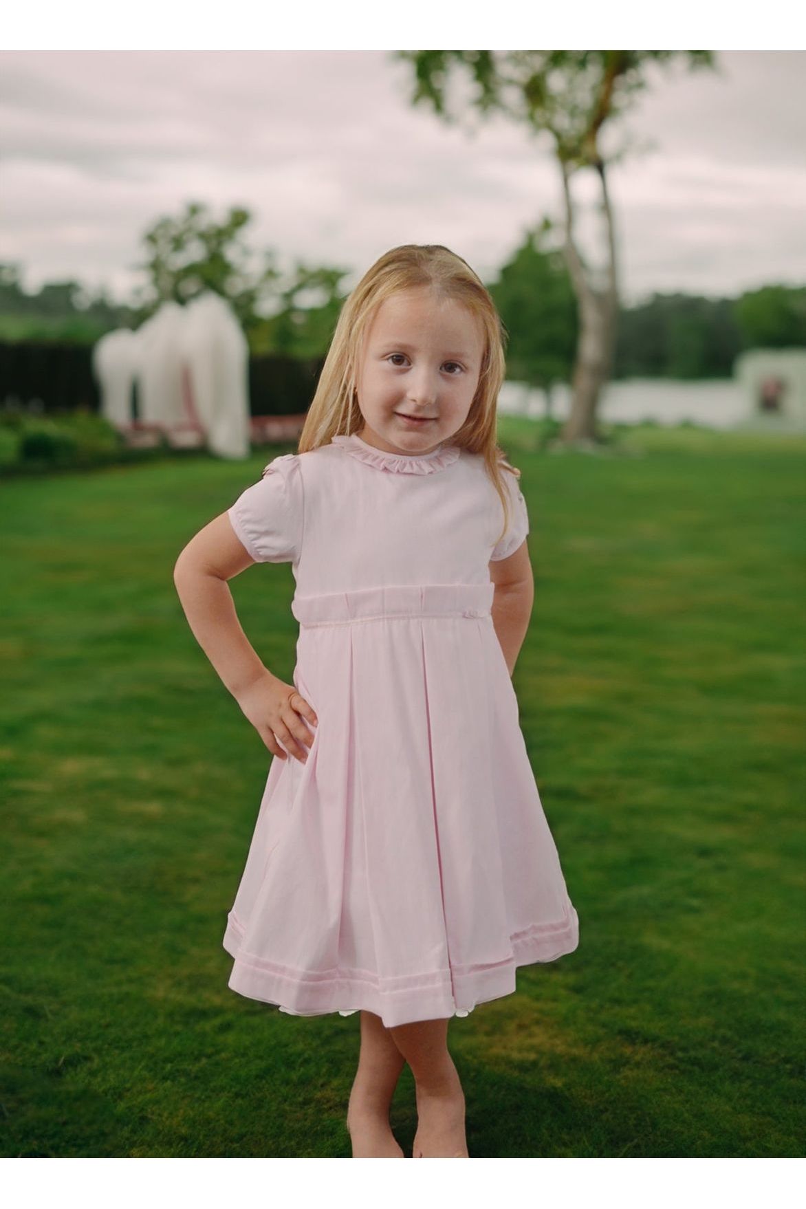 Carriage Boutique pink pique baby girl dress with ruffled collar, worn by a young girl in a grassy outdoor setting.