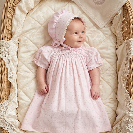 Carriage Boutique baby girl wearing a pink cotton dress with rosebud print and matching lace-trimmed bonnet in a basket