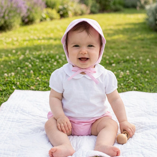 Carriage Boutique baby girl wearing a pink seersucker sun hat and white shirt sitting on a blanket in a garden