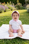Carriage Boutique baby girl wearing a pink seersucker sun hat and white shirt sitting on a blanket in a garden