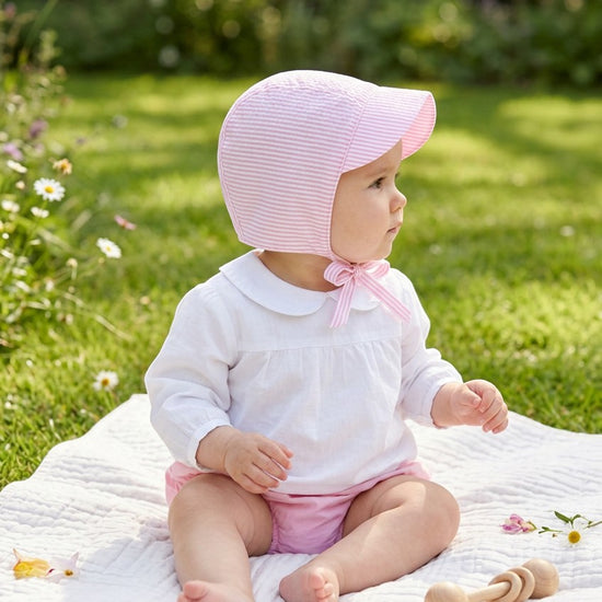 Carriage Boutique baby girl pink seersucker sun hat with chin tie worn by a baby sitting on a blanket in a garden