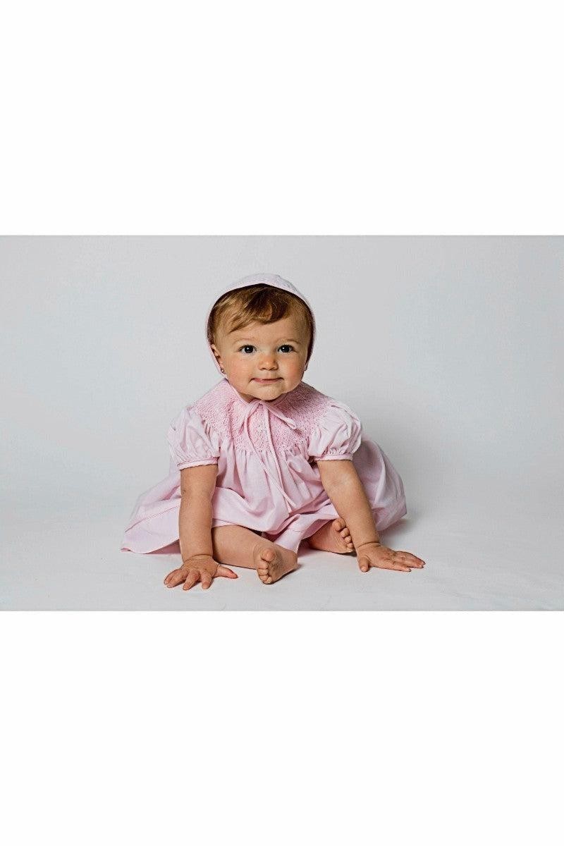 Carriage Boutique pink hand-smocked bishop dress with pearl cross detail and matching bonnet on baby girl.