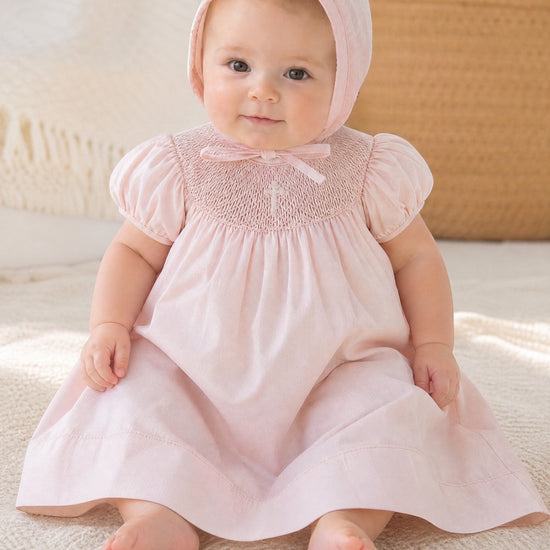 Carriage Boutique pink hand-smocked bishop dress with pearl cross detail and matching bonnet on baby girl.