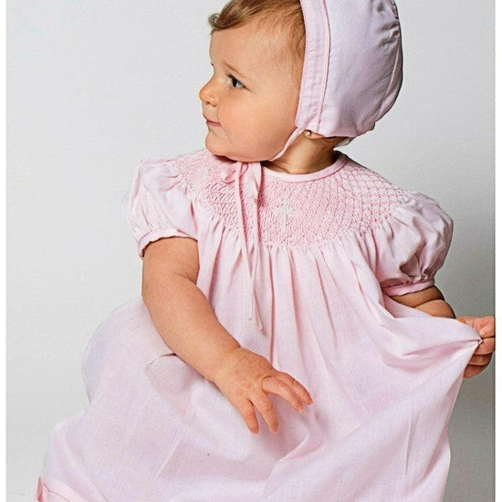 Carriage Boutique pink hand-smocked bishop dress with pearl cross detail and matching bonnet on baby girl.