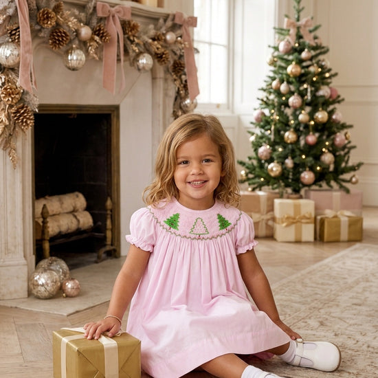 Carriage Boutique toddler girl wearing a light pink cotton bishop dress with hand-smocked green Christmas tree details.