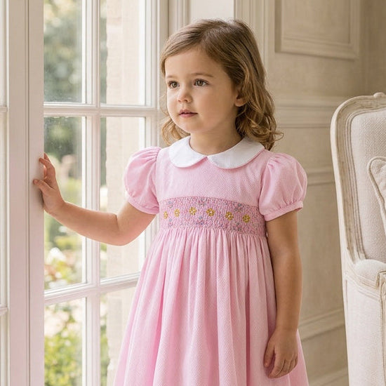 Carriage Boutique pink cotton toddler dress with white collar and smocked yellow flower embroidery by a window