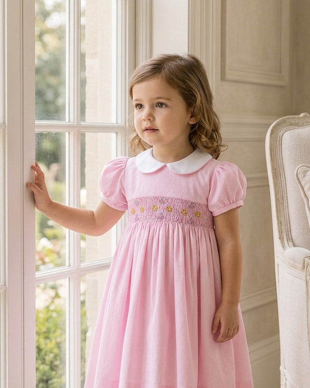 Carriage Boutique pink cotton toddler dress with white collar and smocked yellow flower embroidery by a window